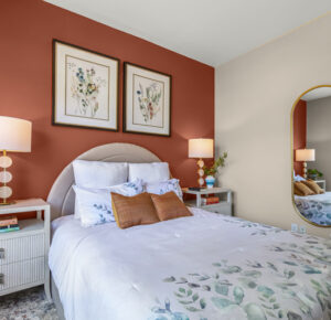 Contemporary - Transitional guest bedroom by Raashi Design with terracotta accent wall, warm neutral upholstered bed, botanical artwork, mirrored accent, and layered linens.