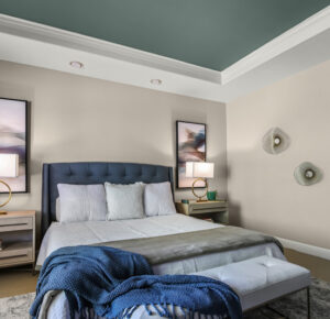Primary bedroom by Raashi Design with blue tray ceiling, dark grey custom upholstered bed with tufted headboard, layered blue and white bedding, oak nightstands, framed art, and sculptural wall art.
