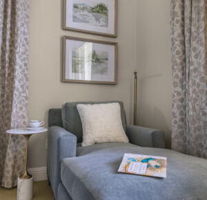 Elegant grey fabric chaise lounge with plush cream pillow, styled in a cozy reading nook with patterned curtains, a modern side table, and two framed nature art prints. Ideal for relaxation and stylish home decor.